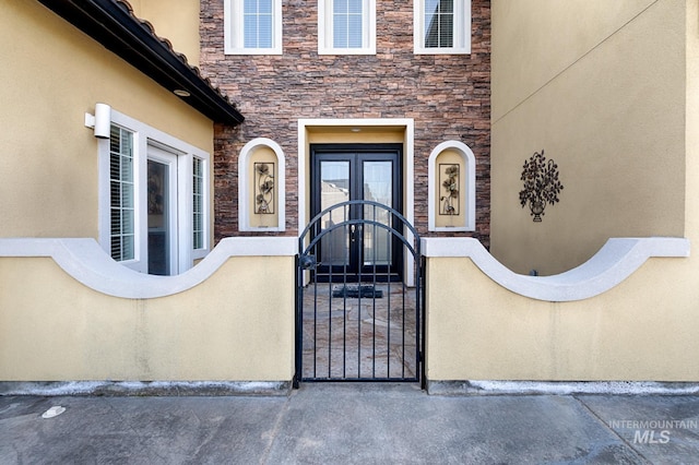 view of exterior entry with stucco siding, a gate, stone siding, fence, and french doors