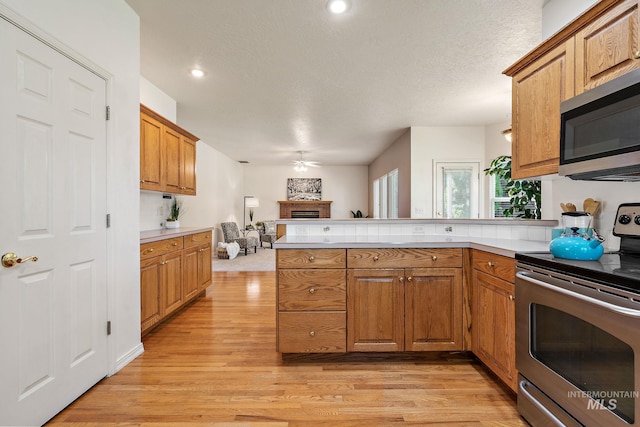 kitchen featuring appliances with stainless steel finishes, a peninsula, ceiling fan, light wood-style floors, and a fireplace