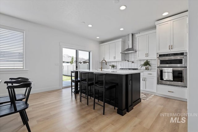 kitchen with backsplash, double oven, a kitchen island with sink, white cabinets, and wall chimney range hood