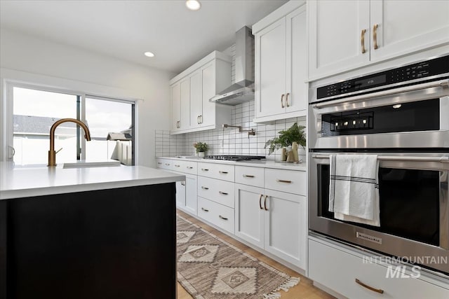 kitchen featuring backsplash, light countertops, stainless steel double oven, wall chimney range hood, and a sink