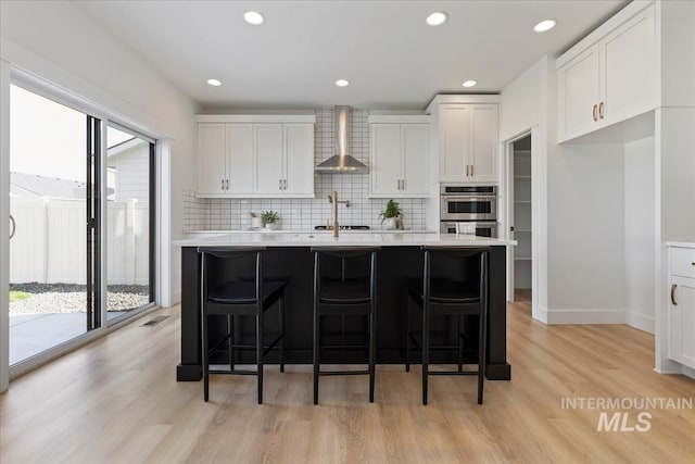 kitchen featuring tasteful backsplash, a center island with sink, light countertops, stainless steel double oven, and wall chimney range hood