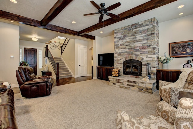 living room with ceiling fan, a stone fireplace, beamed ceiling, and carpet