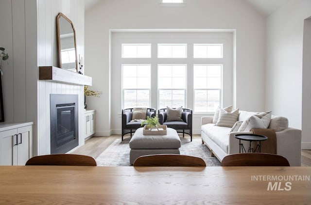 living room featuring light wood finished floors, baseboards, high vaulted ceiling, and a glass covered fireplace