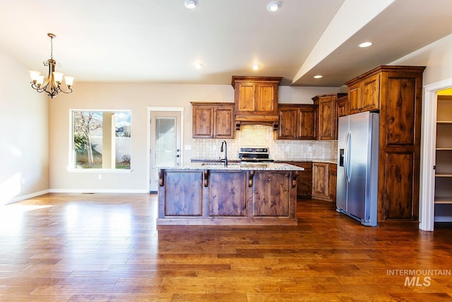 kitchen featuring stainless steel fridge with ice dispenser, light stone counters, a chandelier, a center island with sink, and range