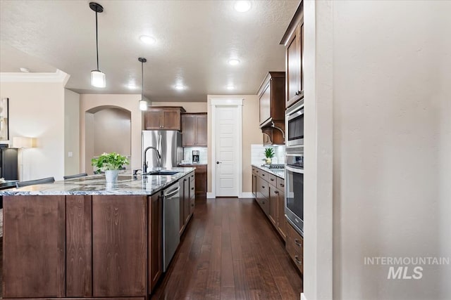 kitchen with dark wood-type flooring, a center island with sink, a sink, appliances with stainless steel finishes, and light stone countertops