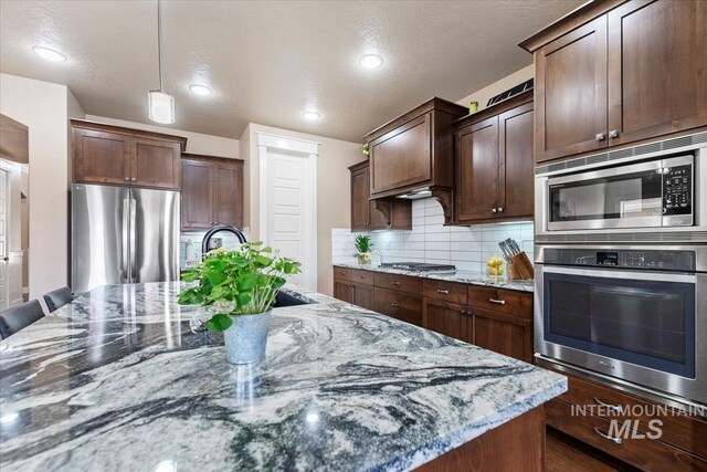 kitchen with light stone counters, a sink, hanging light fixtures, appliances with stainless steel finishes, and backsplash