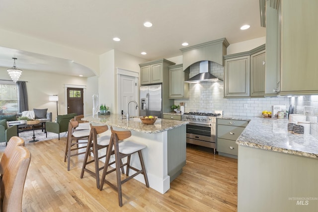 kitchen with wall chimney range hood, light wood-style floors, a breakfast bar area, arched walkways, and stainless steel appliances