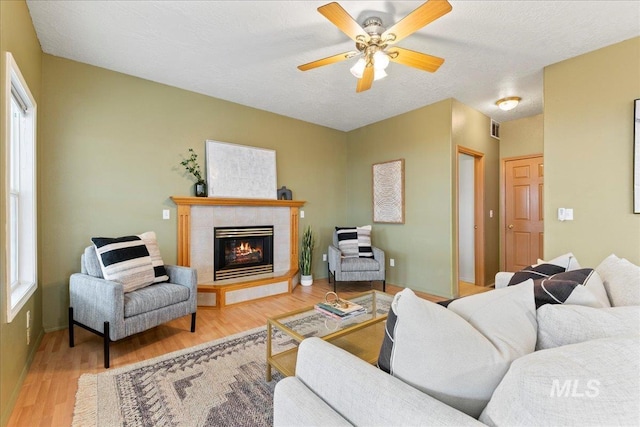 living room with a tiled fireplace, ceiling fan, a textured ceiling, and light hardwood / wood-style floors