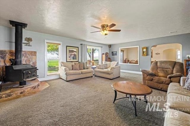 carpeted living room featuring arched walkways, a ceiling fan, a wood stove, a textured ceiling, and baseboards