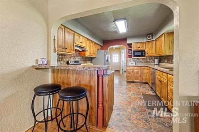 kitchen with black microwave, arched walkways, under cabinet range hood, a peninsula, and freestanding refrigerator