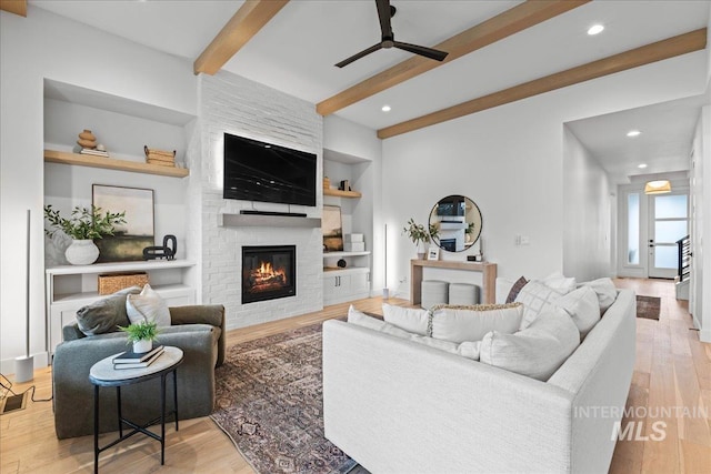 living area with built in shelves, beam ceiling, light wood-style flooring, a large fireplace, and ceiling fan