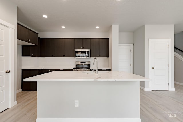 kitchen with an island with sink, appliances with stainless steel finishes, dark brown cabinets, and light hardwood / wood-style flooring