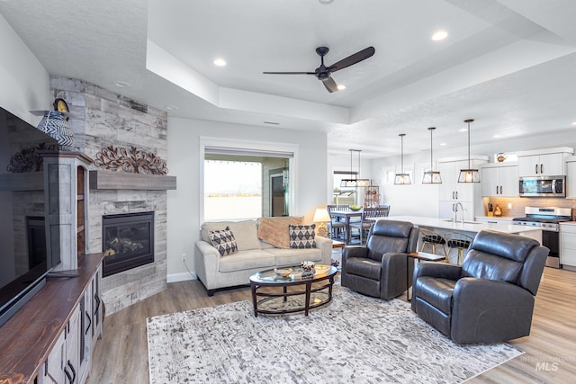 living room featuring ceiling fan, sink, light hardwood / wood-style flooring, a tray ceiling, and a tiled fireplace