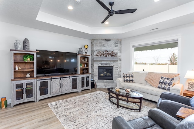 living room featuring ceiling fan, a stone fireplace, light hardwood / wood-style flooring, and a tray ceiling