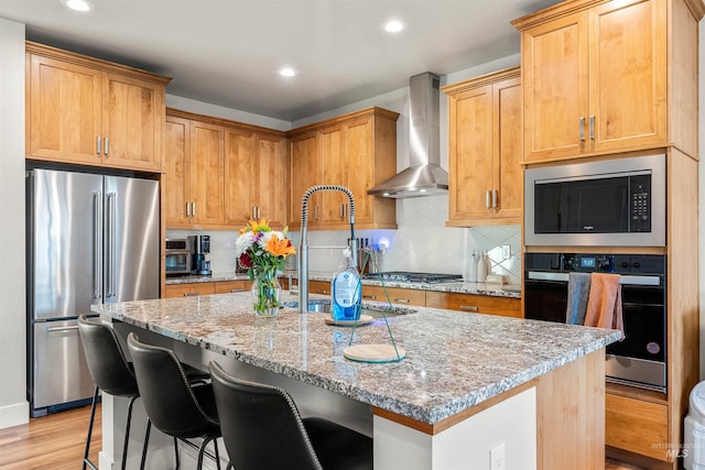 kitchen with light hardwood / wood-style flooring, light stone counters, a kitchen island with sink, wall chimney range hood, and stainless steel appliances