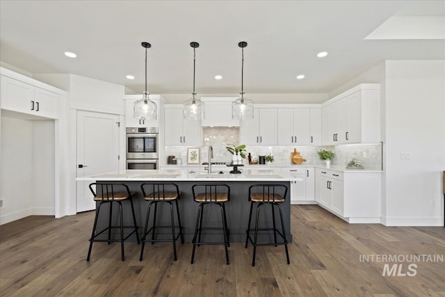 kitchen with white cabinetry, backsplash, an island with sink, and double oven