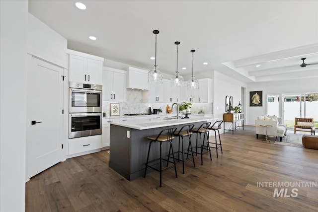 kitchen featuring dark hardwood / wood-style flooring, an island with sink, pendant lighting, stainless steel double oven, and white cabinets