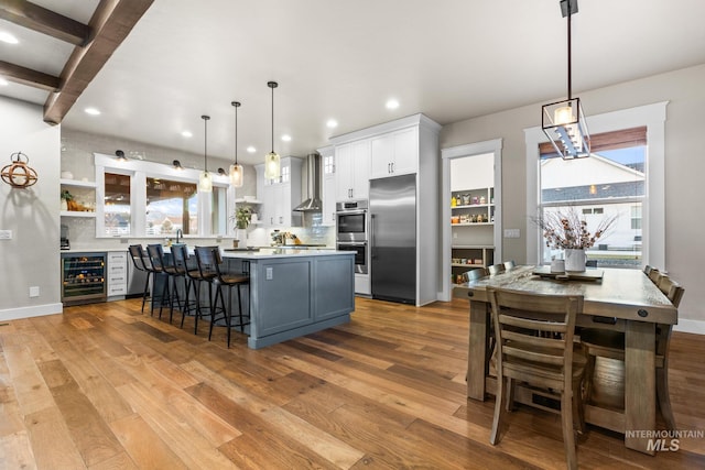 kitchen featuring wine cooler, light countertops, appliances with stainless steel finishes, light wood-style floors, and wall chimney exhaust hood