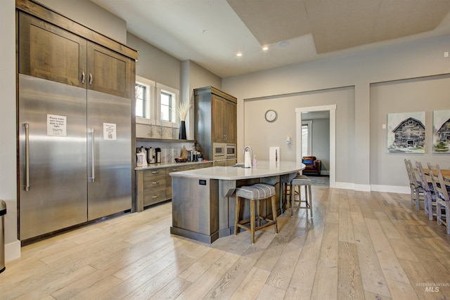 kitchen with built in appliances, a kitchen island with sink, a sink, light countertops, and light wood-type flooring