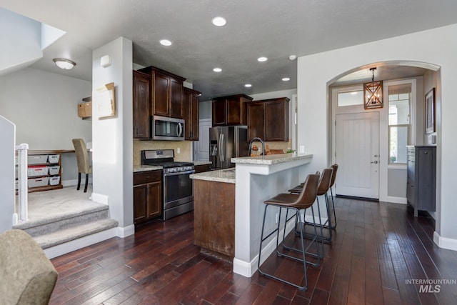kitchen featuring stainless steel appliances, arched walkways, a peninsula, dark brown cabinetry, and dark wood-style flooring