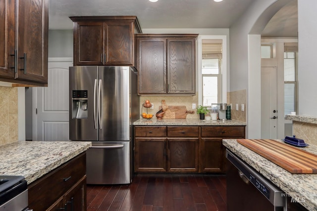 kitchen with stainless steel appliances, arched walkways, tasteful backsplash, light stone counters, and dark brown cabinetry