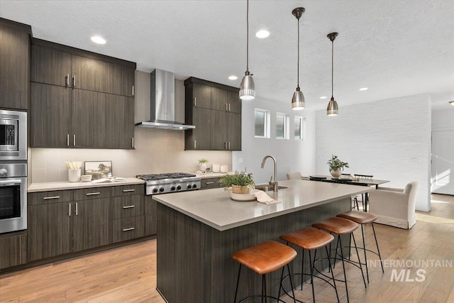 kitchen featuring a breakfast bar, a sink, stainless steel appliances, light wood-style floors, and wall chimney range hood