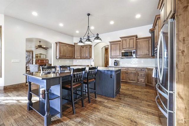 kitchen with tasteful backsplash, appliances with stainless steel finishes, ceiling fan, arched walkways, and a kitchen island with sink