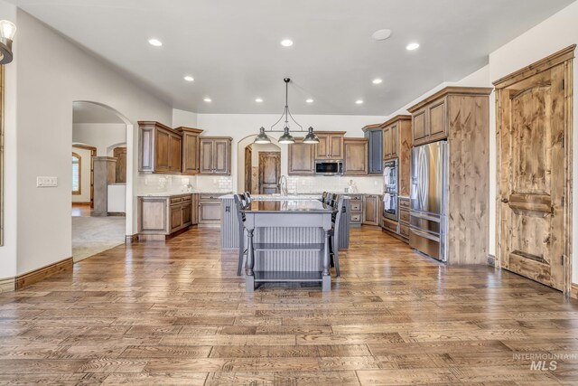 kitchen featuring dark wood-style floors, stainless steel appliances, a kitchen island with sink, and arched walkways