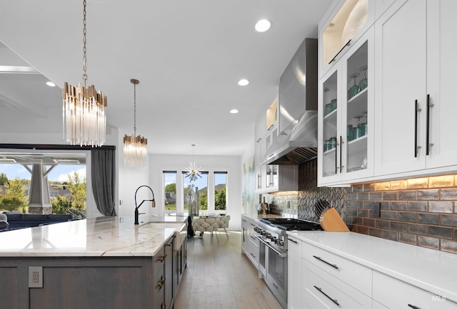 kitchen featuring white cabinetry, wall chimney exhaust hood, tasteful backsplash, range with two ovens, and a center island with sink