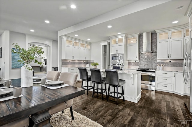 kitchen featuring white cabinets, appliances with stainless steel finishes, wall chimney exhaust hood, dark hardwood / wood-style floors, and a center island with sink