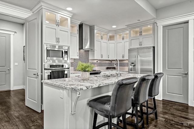 kitchen featuring appliances with stainless steel finishes, wall chimney exhaust hood, sink, a kitchen island with sink, and light stone counters