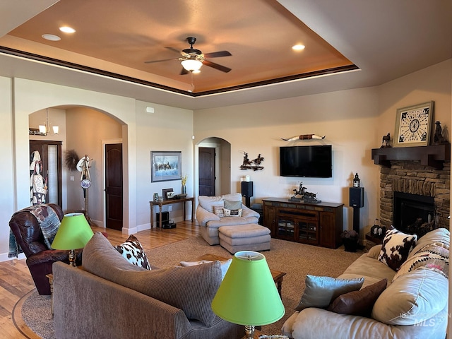 living room featuring ceiling fan, a stone fireplace, light wood-type flooring, and a tray ceiling