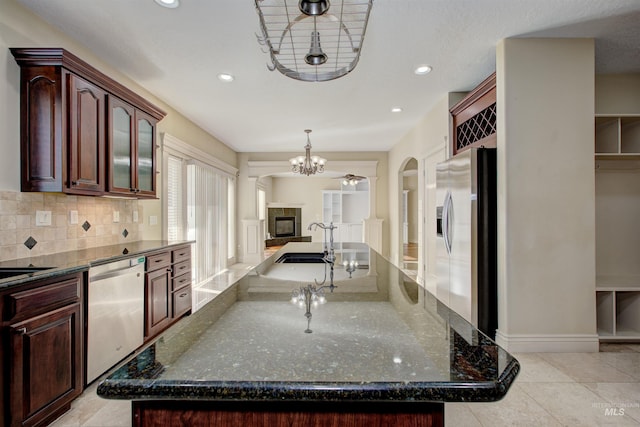 kitchen featuring decorative light fixtures, stainless steel appliances, an island with sink, backsplash, and dark stone counters