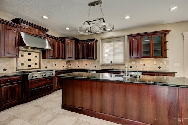 kitchen with stainless steel gas stovetop, decorative light fixtures, dark stone countertops, and a center island with sink