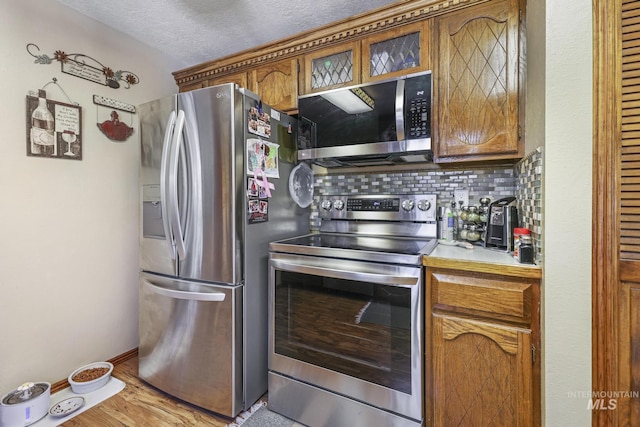 kitchen featuring light countertops, appliances with stainless steel finishes, brown cabinets, decorative backsplash, and glass insert cabinets