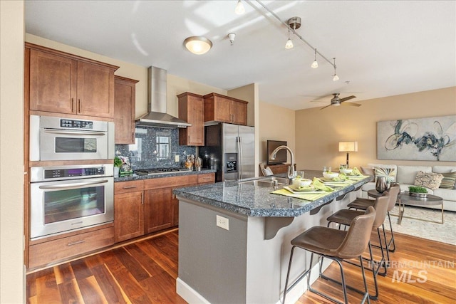 kitchen with wall chimney exhaust hood, appliances with stainless steel finishes, dark wood-type flooring, and a sink