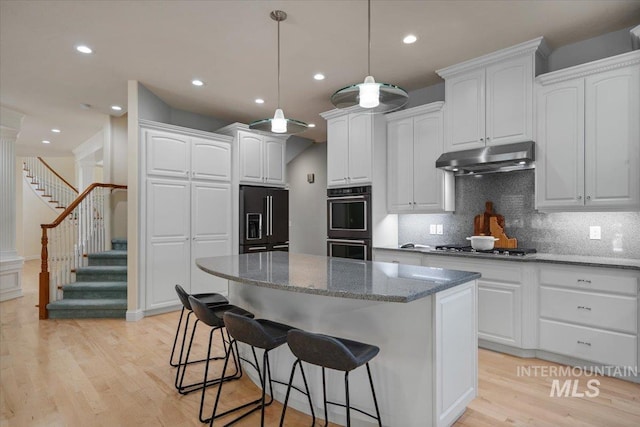 kitchen featuring high end black refrigerator, multiple ovens, under cabinet range hood, light wood-style floors, and a kitchen bar