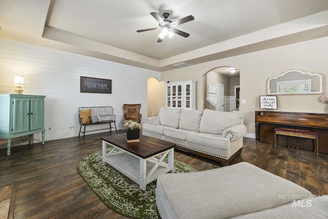 living area featuring arched walkways, a tray ceiling, a ceiling fan, dark wood finished floors, and wooden walls