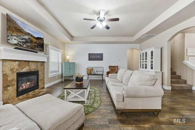 living area featuring a tray ceiling, ceiling fan, arched walkways, a tile fireplace, and baseboards