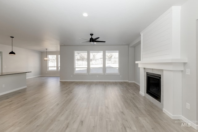 unfurnished living room with ceiling fan with notable chandelier, a tile fireplace, baseboards, and light wood-style floors