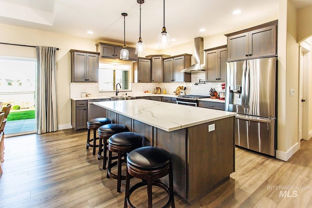 kitchen featuring wall chimney range hood, light wood-type flooring, a kitchen island, decorative light fixtures, and appliances with stainless steel finishes