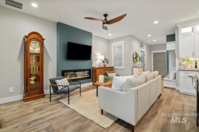 living room featuring ceiling fan, light wood-style floors, recessed lighting, a glass covered fireplace, and baseboards