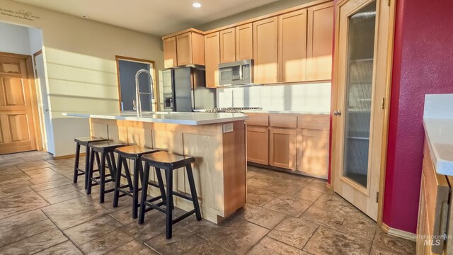 kitchen with stainless steel microwave, light countertops, black fridge with ice dispenser, a kitchen island with sink, and light brown cabinetry