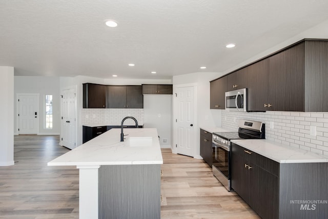 kitchen with stainless steel appliances, light wood-style floors, a kitchen island with sink, a sink, and dark brown cabinets