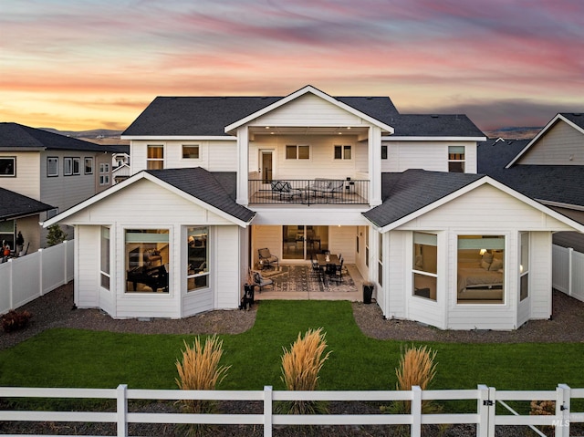 back house at dusk with a patio, a lawn, and a balcony