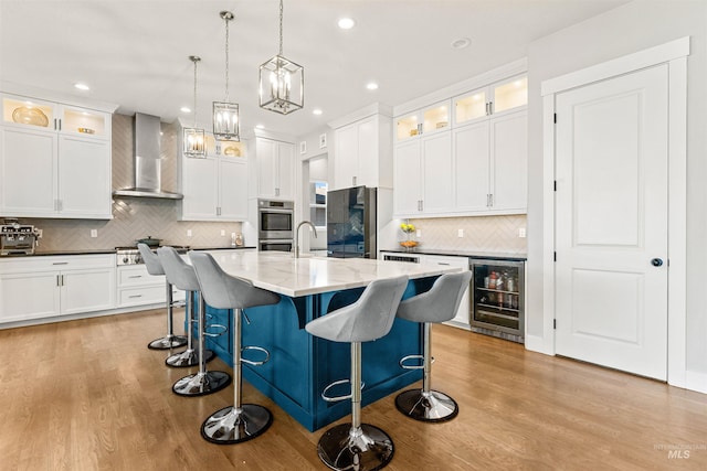 kitchen with beverage cooler, wall chimney range hood, stainless steel appliances, hanging light fixtures, and a kitchen island with sink