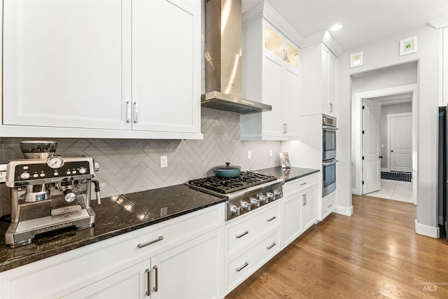 kitchen with dark stone countertops, appliances with stainless steel finishes, light hardwood / wood-style flooring, wall chimney exhaust hood, and white cabinets