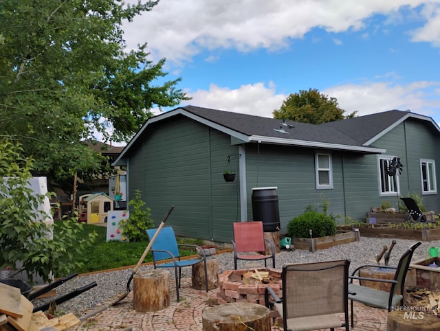 back of property with a fire pit, a patio area, a shingled roof, and a vegetable garden