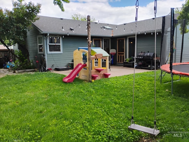 rear view of house featuring a trampoline, a patio, a lawn, and a shingled roof