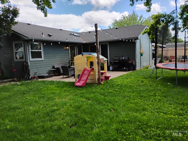 rear view of house featuring a trampoline, a patio, a yard, and a shingled roof
